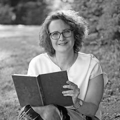 Heike Hamboch Portrait zeigt die Autorin vor einem ländlichen Hintergrund mit einem Buch in der Hand. Sie lächelt in die Kamera.