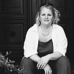 Das Portrait von Nadine Dela zeigt eine Frau vor einer Tür sitzend und in die Kamera lächelnd