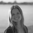 Die Autorin Sascha Sprikut lächelt in die Kamera. Hinter ihr kann man das Meer und einen Strand sehen.