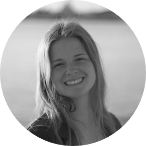 Die Autorin Sascha Sprikut lächelt in die Kamera. Hinter ihr kann man das Meer und einen Strand sehen.